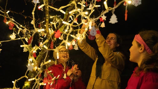 Woman and two children hanging baubles on a tree covered in fairy lights.